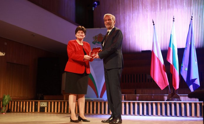 Kobieta gratuluje mężczyźnie na scenie podczas uroczystości, w tle flagi Polski, Bułgarii i UE
