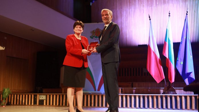 Kobieta gratuluje mężczyźnie na scenie podczas uroczystości, w tle flagi Polski, Bułgarii i UE
